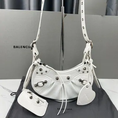 A white studded shoulder bag with a buckled strap, hanging in front of a shop sign with its dust bag below
