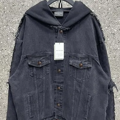 A washed grey denim jacket laid flat on concrete, chest pockets buttoned and a paper tag hanging from one sleeve